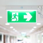 A arrow light box sign of emergency fire exit is hung on the ceiling in hospital walkway, idea for event fire or evacuation drills.
